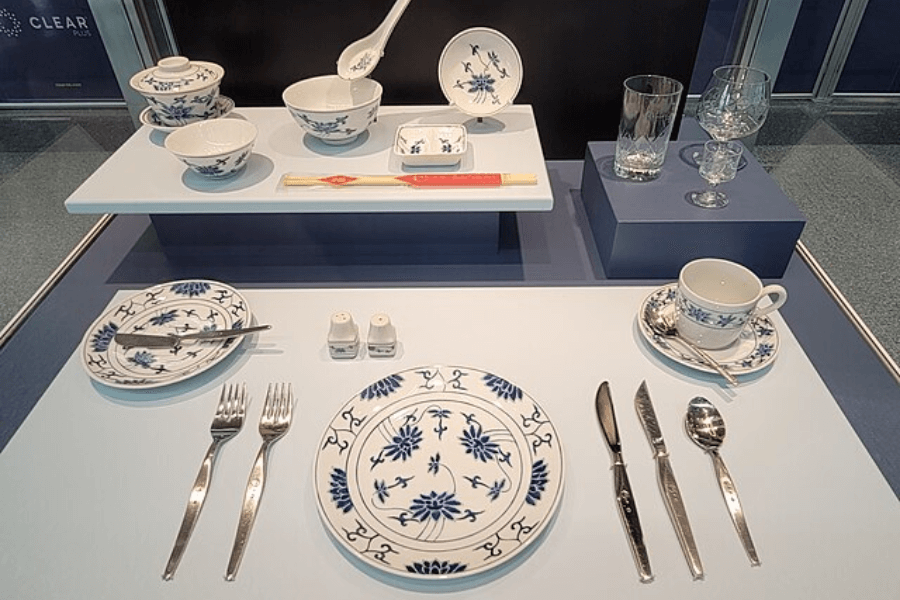 A neatly arranged dining setup featuring elegant blue and white porcelain dishes, bowls, and cups on a display table. The set includes Western-style cutlery, chopsticks in a paper sleeve, salt and pepper shakers, and glassware for different beverages. The arrangement is divided into sections, showcasing different elements of a formal dining experience.