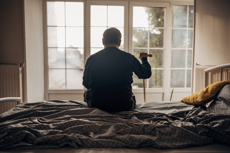 An elderly man sits on the edge of a bed, facing a large window with soft natural light streaming in. He holds a wooden cane in one hand and appears to be deep in thought. The bed is unmade, with gray sheets and a yellow pillow resting against the headboard.