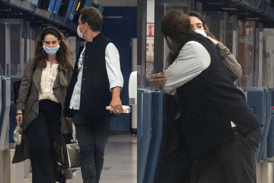 A side-by-side collage of two candid airport photos featuring a man and a woman wearing face masks. In the left image, the woman, dressed in a long coat and carrying shopping bags, walks alongside the man, who wears a black vest over a white shirt and holds a water bottle. In the right image, they are seen leaning against a check-in counter as the man embraces the woman closely, while she tilts her head toward him.