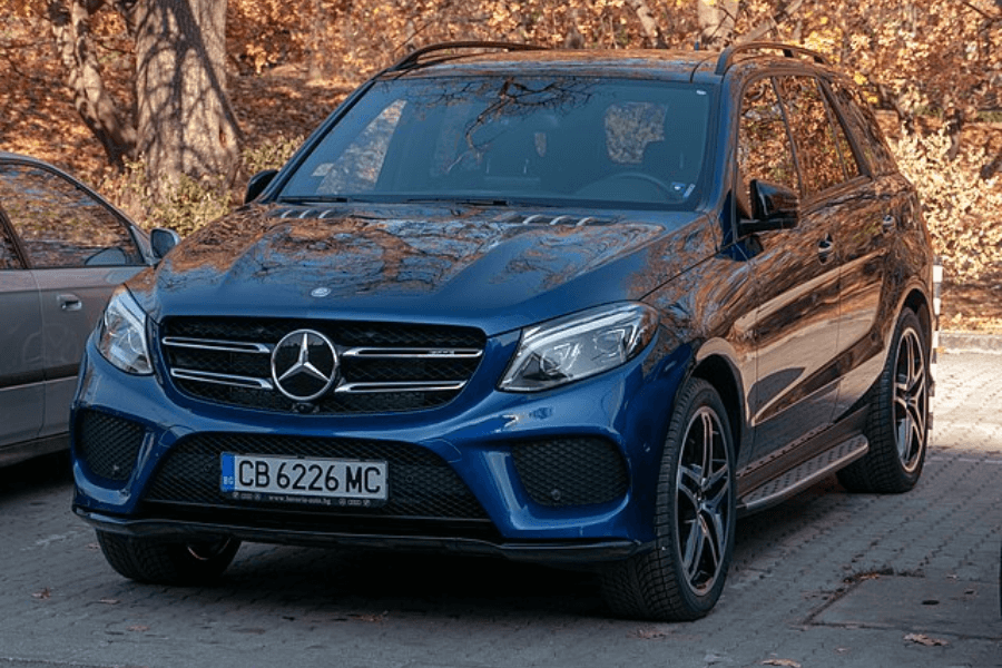 A shiny blue Mercedes-Benz SUV parked on a paved lot with autumn-colored trees in the background. The car features a sleek front grille with the Mercedes emblem, LED headlights, and black alloy wheels. The license plate reads "CB 6226 MC," indicating European registration.