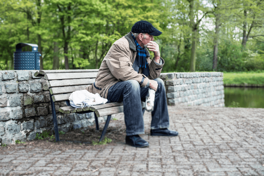 An elderly man wearing a black flat cap, a brown jacket, and a blue plaid scarf sits alone on a wooden bench in a park. He rests his chin on his hand, gazing into the distance with a thoughtful expression. A small plastic bag and a piece of cloth are beside him, while a stone wall and a pond with lush green trees surround the scene.