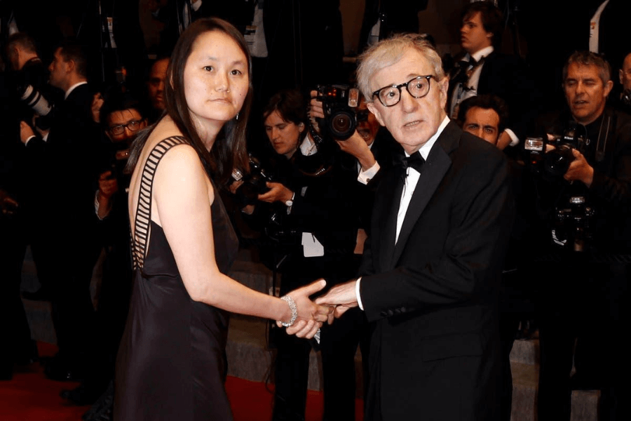 A couple poses on the red carpet at a formal event, surrounded by photographers and flashing cameras. The woman wears a black evening gown with an intricate strap design on the shoulder, and a silver bracelet. The man, dressed in a classic black tuxedo with a bow tie and round glasses, looks slightly surprised as they hold hands. The background is filled with photographers and event attendees in black-tie attire.