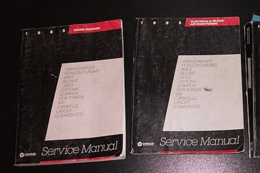 Two worn 1985 Chrysler service manuals with black covers and red headers are placed side by side on a dark surface. The left manual is labeled "Wiring Diagrams," while the right one covers "Electrical & Heater Air Conditioning." Both list various car models, including Omni Charger, Horizon Turismo, Aries, Reliant, Laser, Daytona, LeBaron, New Yorker, and more.