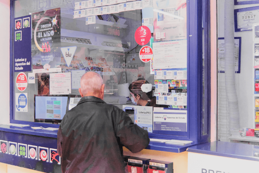 An elderly man in a black leather jacket stands at a lottery ticket counter inside a shop. The glass window is decorated with colorful posters, signs, and jackpot amounts for various lottery games. A masked cashier is visible behind the counter, assisting customers in purchasing tickets.