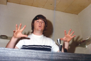 A young man with shaggy brown hair raises both hands expressively while standing in a recording studio. He wears a white t-shirt with navy blue stripes, and a microphone is positioned in front of him. The perforated soundproofing walls and focused expression suggest he is in the middle of a passionate musical performance or production session.