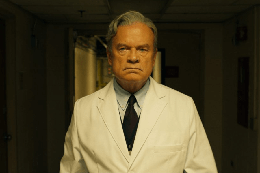 A stern-looking older man with slicked-back gray hair wears a white lab coat over a dress shirt and dark tie, standing in a dimly lit hallway. His intense gaze and furrowed brow give him an authoritative and serious demeanor. The background features a slightly worn hospital or laboratory corridor with exposed wiring and muted lighting, adding a dramatic and mysterious tone.