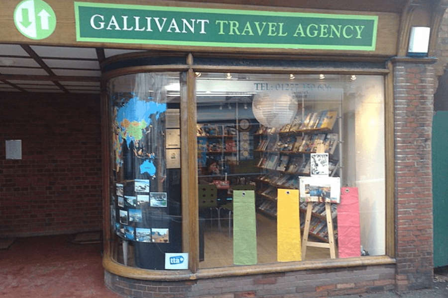 The exterior of "Gallivant Travel Agency," a small travel office with a curved glass window displaying a world map and travel photos. Inside, bookshelves filled with travel guides and brochures are visible, along with a desk where a staff member is seated. A wooden easel with promotional materials stands in the window.