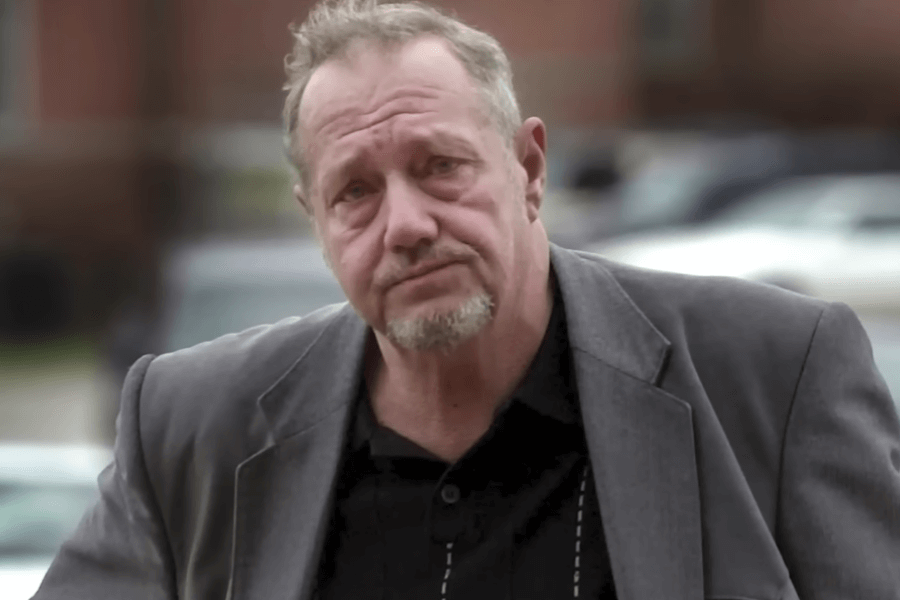 A middle-aged man with short, slightly disheveled hair and a goatee stands outdoors with a serious expression. He wears a dark gray blazer over a black shirt, with glasses hanging from his collar. The blurred background features parked cars and a brick building, suggesting an urban or residential setting.