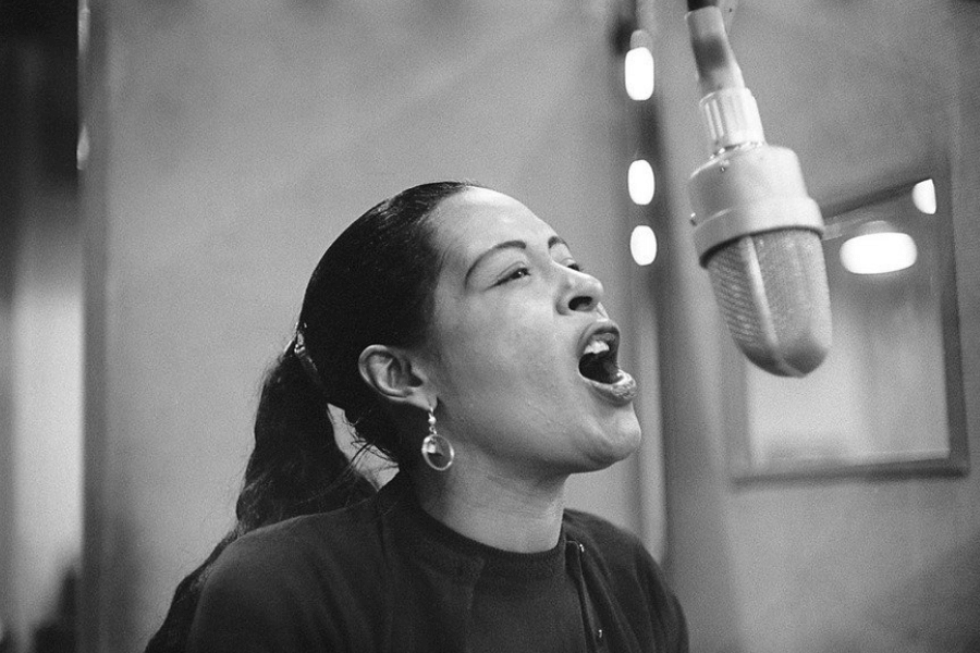 A black-and-white photograph captures a female jazz singer mid-performance, passionately singing into a large vintage studio microphone. Her hair is pulled back, and she wears elegant drop earrings and a dark outfit, with emotion radiating from her open mouth and expressive face. The soft lighting and blurred background create a timeless, intimate atmosphere.