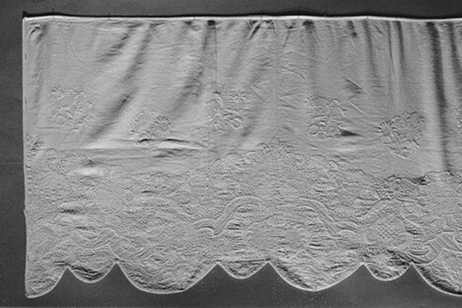 A black-and-white image of an intricately embroidered fabric panel featuring floral patterns and scalloped edges. The lower section showcases detailed lace-like embroidery, possibly depicting a traditional or decorative textile piece.