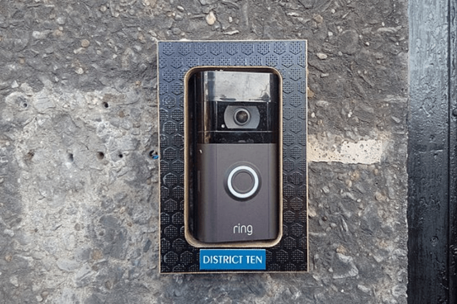 A Ring video doorbell mounted on a textured concrete wall, encased in a decorative black and gold frame labeled "DISTRICT TEN." The doorbell features a camera at the top and an illuminated circular button in the center.