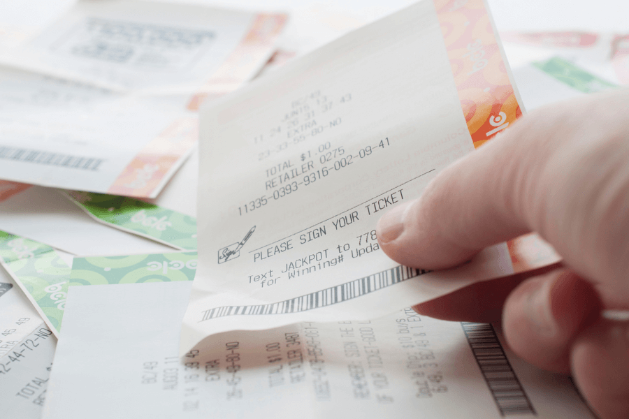 A close-up of a person's hand holding a lottery ticket with a barcode and a "Please Sign Your Ticket" message printed on it. Several other lottery tickets are scattered in the background, featuring colorful designs and printed numbers. The image suggests anticipation and the hope of winning.