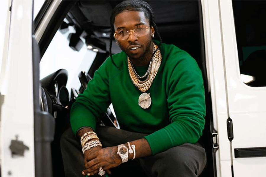 A stylish man with braided hair and a confident expression sits in the open door of a white luxury car. He wears a green sweatshirt, dark pants, and multiple gold chains, rings, and a diamond-encrusted watch. His tinted glasses and relaxed pose add to the image's sleek and powerful aesthetic.