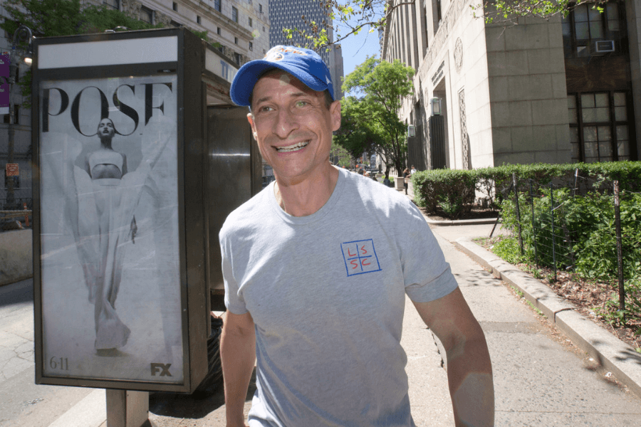 A man wearing a gray T-shirt with a logo and a blue baseball cap smiles while walking down a sunny city sidewalk. Behind him, a bus stop advertisement for the FX series "POSE" features a striking black-and-white image of a person in a dramatic pose. The background includes tall buildings, green trees, and a fenced-in area along the sidewalk.