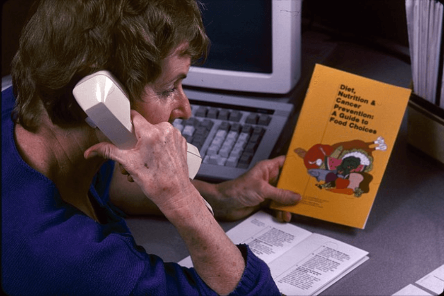 A woman in a blue sweater sits at a desk holding a beige corded telephone to her ear while reading a yellow booklet titled Diet, Nutrition & Cancer Prevention: A Guide to Food Choices. An old-fashioned computer keyboard and monitor are visible in the background, along with paperwork spread out on the desk.