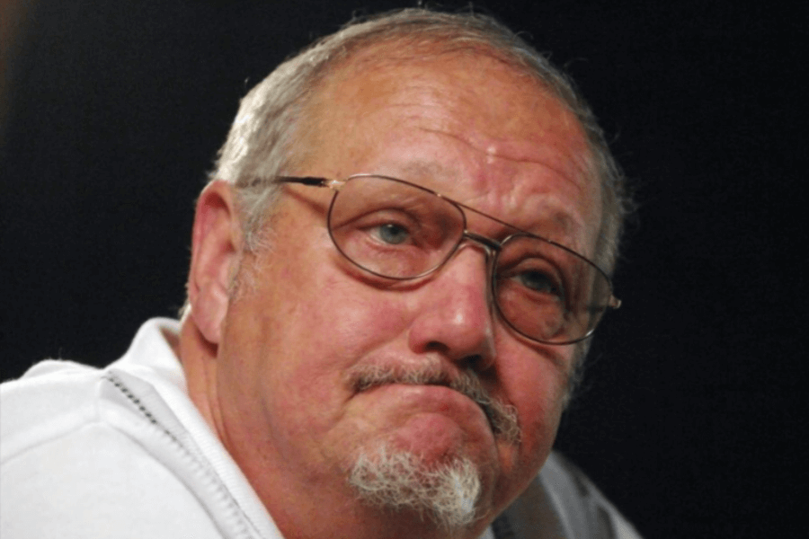 A close-up of an older man with glasses and a short white beard, wearing a white shirt. He has a solemn expression with slightly furrowed brows and lips pressed together, appearing deep in thought. The background is dark and out of focus, emphasizing his face and expression.