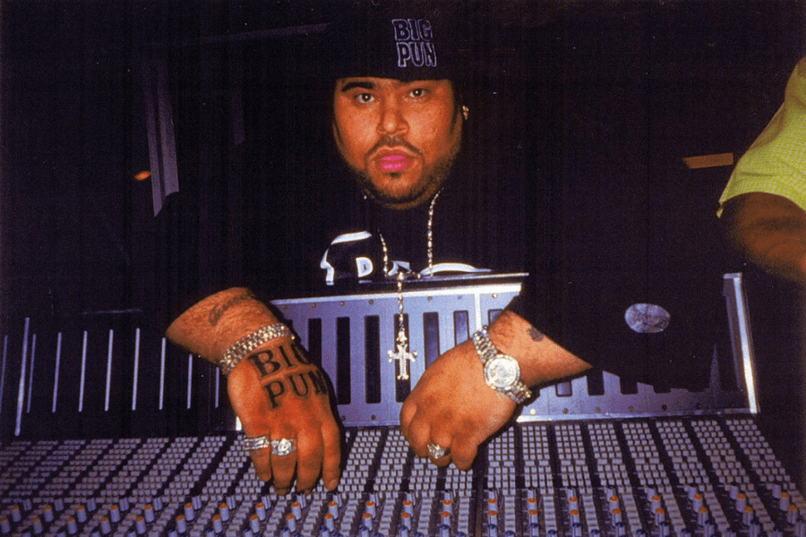 A man wearing a black cap with "BIG PUN" embroidered on it leans over a large mixing console in a dimly lit recording studio. He sports a black t-shirt, multiple gold chains with a cross pendant, and several rings and bracelets, including a tattoo on his hand that reads "BIG PUN." His intense expression and the professional studio setting emphasize his presence in the music industry.