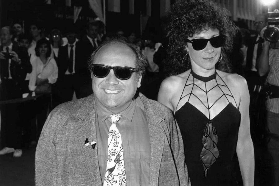 A black-and-white photograph of a smiling man in a suit and a patterned tie, wearing dark sunglasses, standing beside a woman with curly hair, also in sunglasses. She wears a bold geometric-designed dress with a halter neckline. The background is filled with a crowd of onlookers and photographers, suggesting a red carpet or public event.