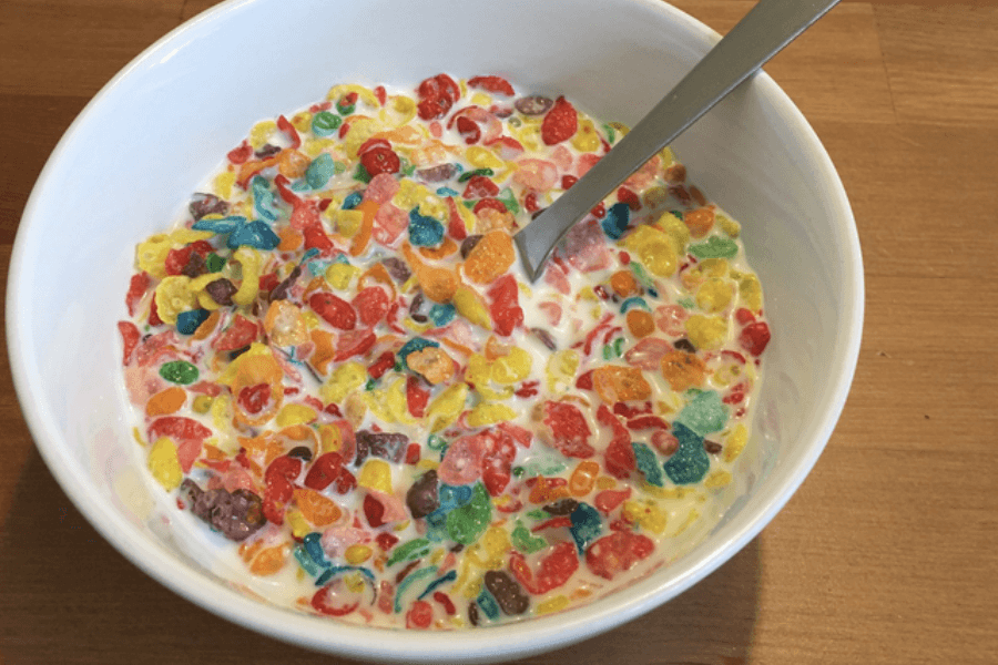 A white bowl filled with colorful, fruit-flavored cereal pieces mixed with milk, creating a slightly soggy texture. A silver spoon is partially submerged in the bowl, resting on the edge, and the bowl sits on a wooden surface.