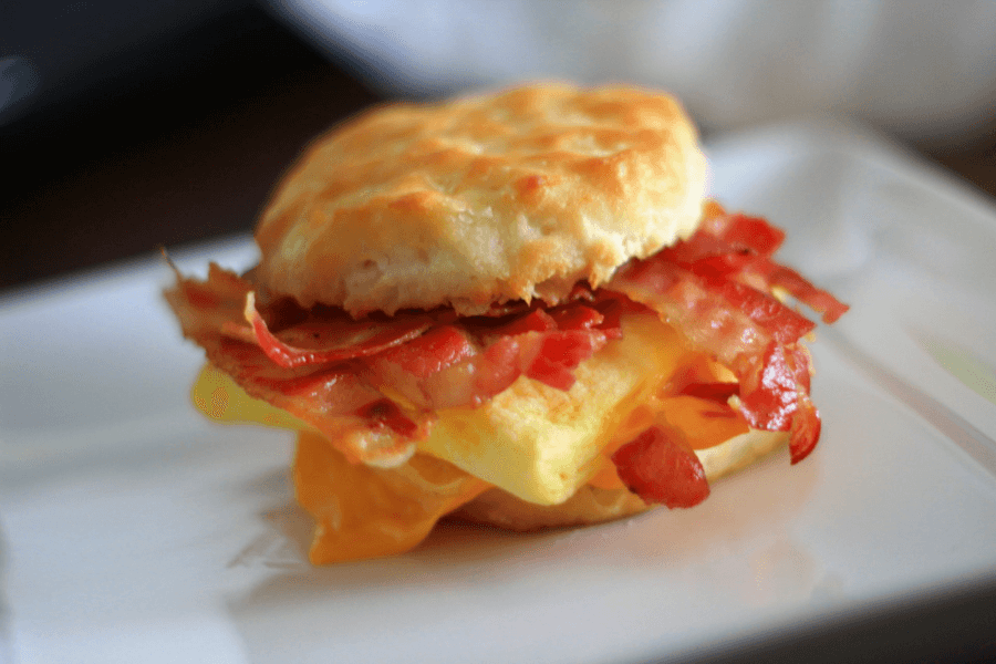 A freshly made breakfast sandwich featuring a golden-brown biscuit, crispy bacon, a fluffy folded egg, and melted cheddar cheese sits on a white plate. The bacon is slightly curled, and the cheese is slightly melting over the egg. The biscuit appears soft and flaky, with a slightly glossy surface.