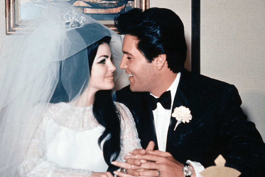 A groom in a black tuxedo with a white boutonniere and a bride in a long-sleeved white wedding dress with a sheer veil sit closely together, gazing into each other's eyes with smiles. The groom clasps the bride’s hands, and they appear joyful. The setting is an indoor wedding reception with a framed painting in the background.