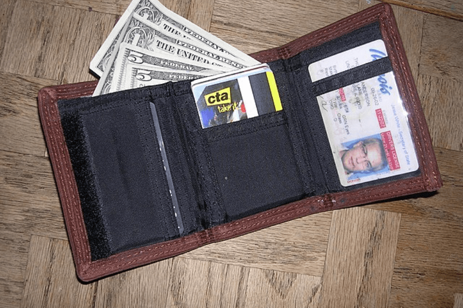 An open brown fabric wallet with black interior compartments placed on a wooden parquet floor. Inside, there are U.S. dollar bills partially sticking out, a CTA transit card, and an identification card with a visible photo.