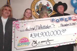 A group of four people stands together holding an oversized Powerball lottery check for $314.9 million, made out to Andrew J. Whittaker Jr. and dated December 26, 2002. The check features the "West Virginia Lottery" logo, and balloons with the same branding are visible in the background. The man in a black cowboy hat and glasses, likely the lottery winner, stands at the podium with a serious expression, while the women beside him smile. The West Virginia state seal is displayed on the wall behind them.