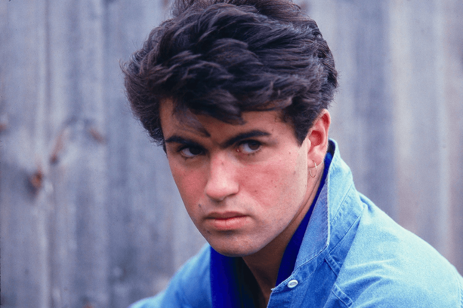 A young man with thick, wavy dark hair and intense eyes gazes off to the side, his expression both serious and captivating. He wears a light blue denim jacket with a dark blue collar and a small gold hoop earring in one ear. The blurred wooden fence in the background adds to the nostalgic and effortlessly cool aesthetic.