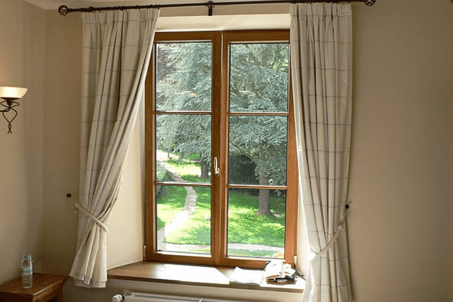 A wooden-framed window with white, checkered curtains tied back, allowing natural light to enter the room. The window overlooks a lush green garden with a winding pathway leading through tall trees. A radiator is positioned below the window, and a wooden side table with a water bottle is visible in the corner.