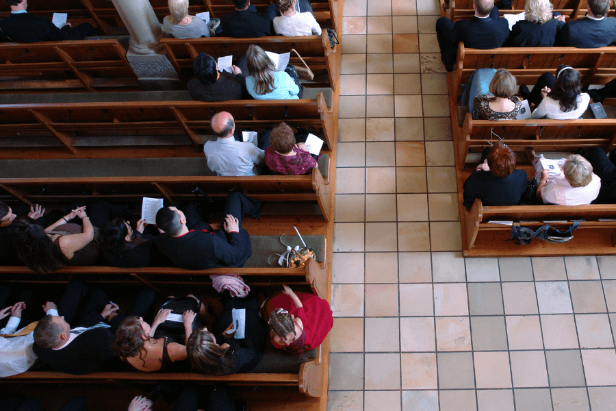 An overhead view of a congregation seated in wooden pews inside a church. The attendees, dressed in formal attire, hold programs or booklets, suggesting they are part of a ceremony or service. A central aisle divides the seating, with a small basket and bouquet placed near the front.