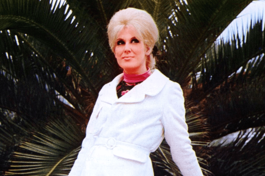 A stylish woman with a voluminous blonde updo poses in front of lush green palm leaves. She wears a chic white coat with a belt cinched at the waist, layered over a high-neck pink and black top. Her dramatic eye makeup and poised expression capture the elegance and glamour of a classic era.