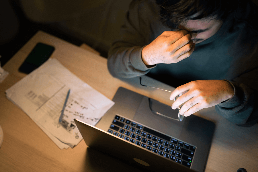 A man in a dark sweatshirt sits at a desk with a laptop, rubbing his eyes while holding his glasses in one hand. Papers with handwritten notes and a pen are scattered nearby, indicating he is working late or feeling stressed. The dim lighting casts shadows, emphasizing his exhaustion.