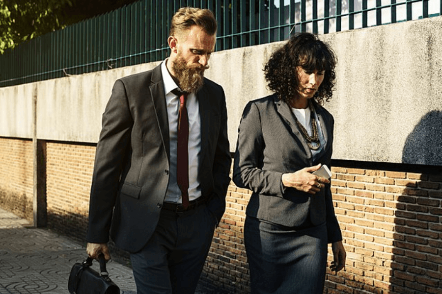 A man and a woman in business attire walk side by side along a sunlit sidewalk near a brick and concrete wall. The man has a beard, wears a dark suit with a red tie, and carries a black leather briefcase. The woman wears a dark gray skirt suit with a white blouse, accessorized with layered necklaces, and holds a smartphone in her hand.