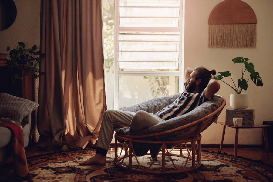 A relaxed man with a beard and tied-back hair lounges in a cozy rattan chair, his hands resting behind his head. Sunlight filters through a window with sheer curtains, casting a warm glow on the bohemian-style living room, which features houseplants, a woven wall hanging, and a patterned rug. A small side table holds a white coffee cup, completing the serene atmosphere.
