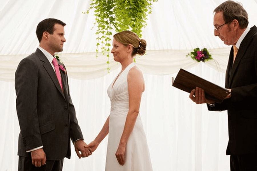 A bride and groom stand inside a white wedding tent, holding hands and gazing at each other as they exchange vows. The groom wears a dark suit with a pink tie and a boutonnière, while the bride wears a white sleeveless gown with her hair in an updo. A wedding officiant in a black suit and yellow tie reads from a book as he conducts the ceremony.