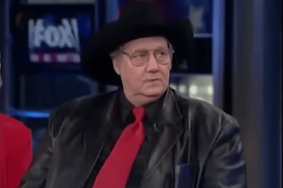 A man in a black cowboy hat, glasses, and a leather jacket with a red tie appears on a Fox News broadcast. He has a serious expression as he looks to the side, seemingly engaged in conversation. The studio's dim lighting and blue-toned background screens create a professional interview setting.