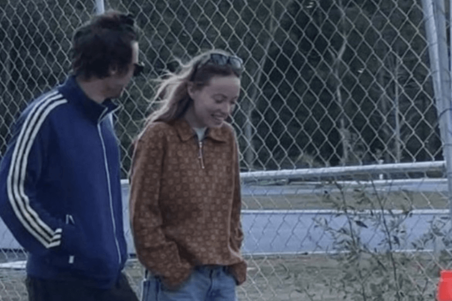 A casually dressed couple walks outdoors near a chain-link fence, smiling and engaged in conversation. The man wears a navy blue Adidas track jacket and sunglasses, while the woman wears a brown zip-up sweater with a patterned design, jeans, and sunglasses resting on her head. The setting appears relaxed, with a road and greenery in the background.