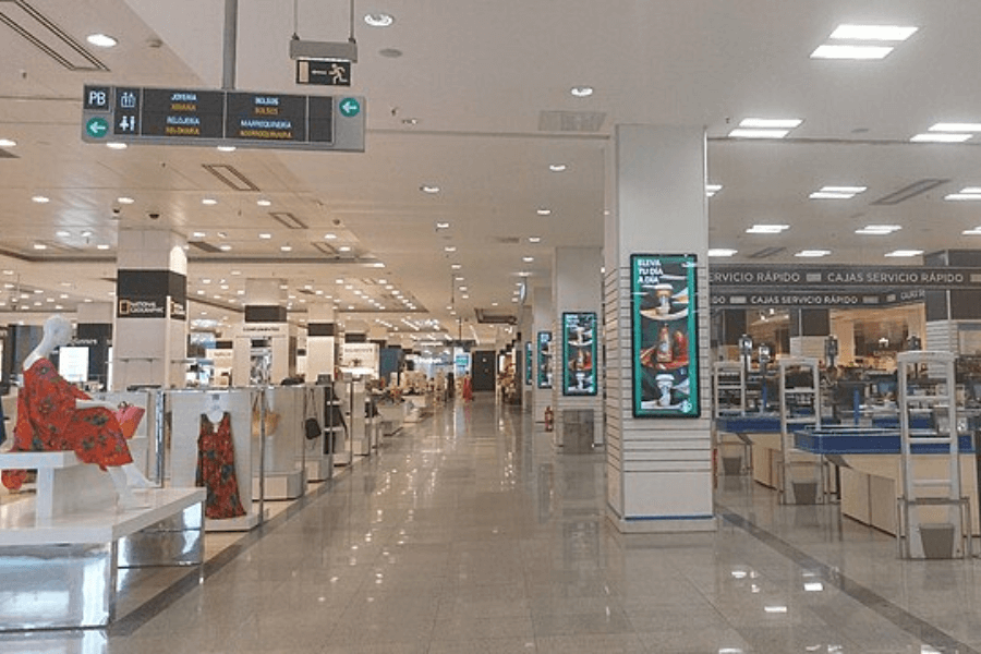 A spacious department store with a polished tile floor and bright overhead lighting. Various clothing and accessory displays are arranged throughout, including mannequins dressed in colorful outfits. In the background, there are checkout counters labeled "Cajas Servicio Rápido" and electronic signboards displaying advertisements.