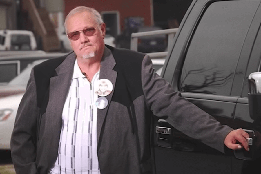 A middle-aged man with glasses and a goatee leans on a black pickup truck, wearing a gray suit jacket over a striped shirt. He has a serious expression and wears two round pins on his jacket. The background features a parking lot with multiple vehicles and a brick building.