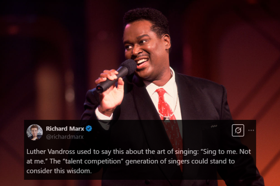 A male singer performs on stage, holding a microphone and smiling warmly. He is dressed in a black suit with a white shirt and a red tie adorned with subtle patterns. The stage lighting casts a warm glow, complementing his engaging and charismatic presence.