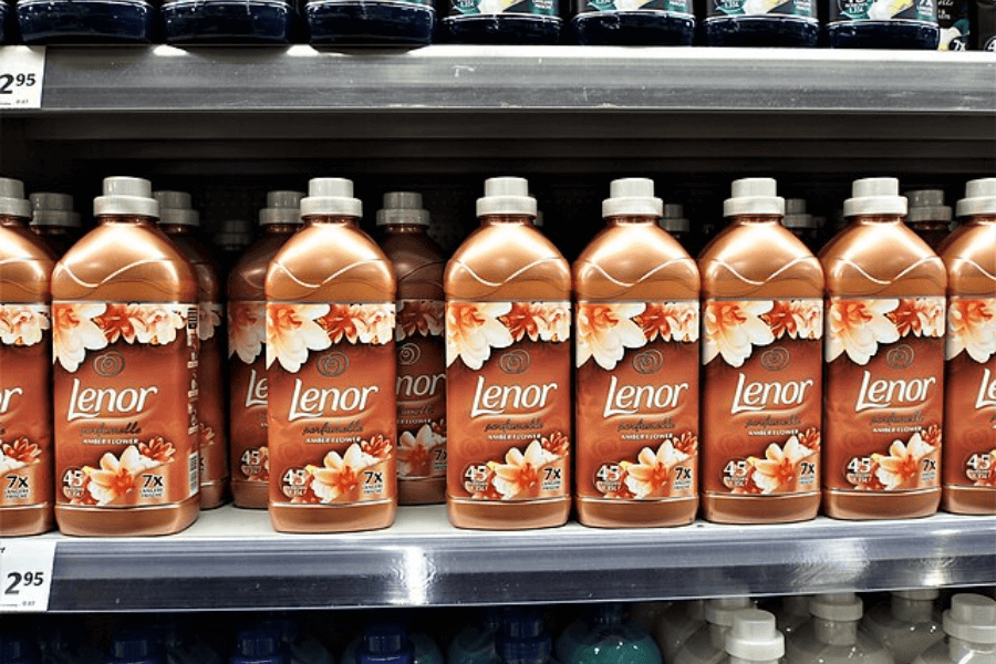 A supermarket shelf stocked with multiple bottles of Lenor fabric softener in an amber and floral scent. The bottles are bronze-colored with white and orange flower illustrations on the label, and a price tag of "2,95" is visible on the shelf.