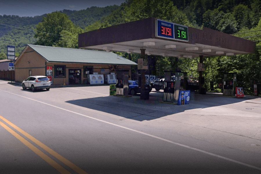 A rural gas station with a brown canopy and multiple fuel pumps is located along a two-lane road, surrounded by lush green hills. The attached convenience store, "C & L," has a green metal roof and displays various signs, coolers, and ice machines outside. A white SUV is parked near the entrance, while other vehicles are at the pumps. The digital gas price sign shows regular gasoline at $3.19 and diesel at $3.99.