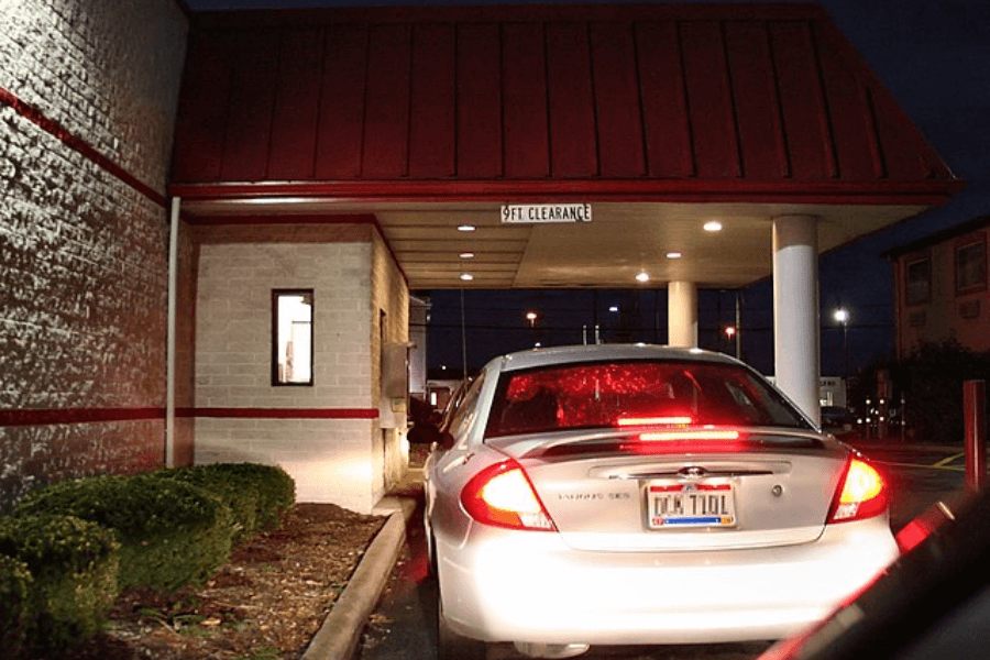 A white Ford Taurus with its brake lights on waits in line at a fast-food drive-thru at night. The restaurant has a red roof and a "9 FT. CLEARANCE" sign above the covered drive-thru lane, with a lit service window visible on the left.