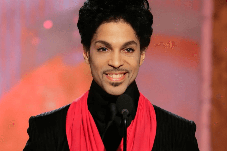 A musician with a stylish, curly updo and a thin mustache smiles while standing at a microphone. He is wearing a black textured suit with a high collar and a vibrant pink scarf draped around his neck. The background features warm-toned, softly blurred lights, giving the scene an elegant and celebratory atmosphere.