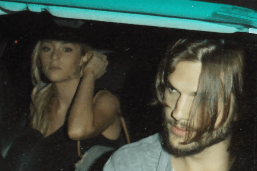 A man with long hair partially covering his face and a beard sits in the front seat of a car at night, looking to the side with a serious expression. Behind him, a blonde woman in a black outfit adjusts her hair while gazing forward with a neutral expression. The dim lighting and reflections on the car window give the image a candid, paparazzi-style feel.