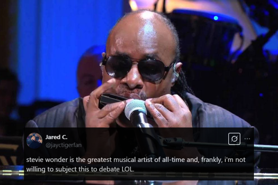 A male musician wearing dark sunglasses and a black suit plays the harmonica while seated at a piano. He leans into the microphone, deeply engrossed in the performance, as stage lighting casts a cool blue glow around him. The blurred background reveals a live band setup, enhancing the concert atmosphere.