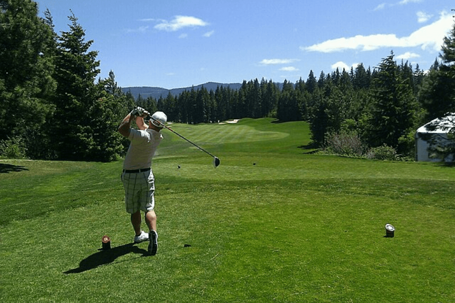 A golfer in a light-colored outfit and plaid shorts swings a driver on a lush green golf course surrounded by tall pine trees. The sky is bright blue with a few scattered clouds, and the fairway extends into the distance with sand bunkers visible.