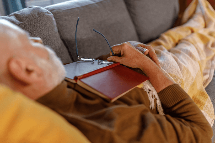 An elderly man with a white beard is resting on a gray couch, covered with a yellow plaid blanket. He is holding a pair of reading glasses in one hand, with a closed red book resting on his chest. His eyes are closed, suggesting he has dozed off while reading.