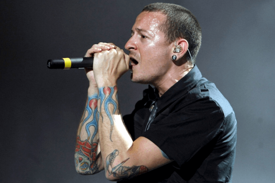 A passionate male singer with a shaved head and in-ear monitors performs on stage, gripping a microphone tightly. He is wearing a black button-up shirt, and his arms are covered in colorful flame and skull tattoos. His face glistens with sweat as he sings intensely against a dark, moody background.