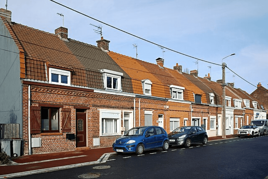 A row of traditional European brick houses with sloped tiled roofs and dormer windows lines a residential street. Several cars, including a blue Citroën, are parked along the curb, and a utility pole with overhead wires runs parallel to the houses. The sky is clear with soft sunlight casting shadows on the buildings.